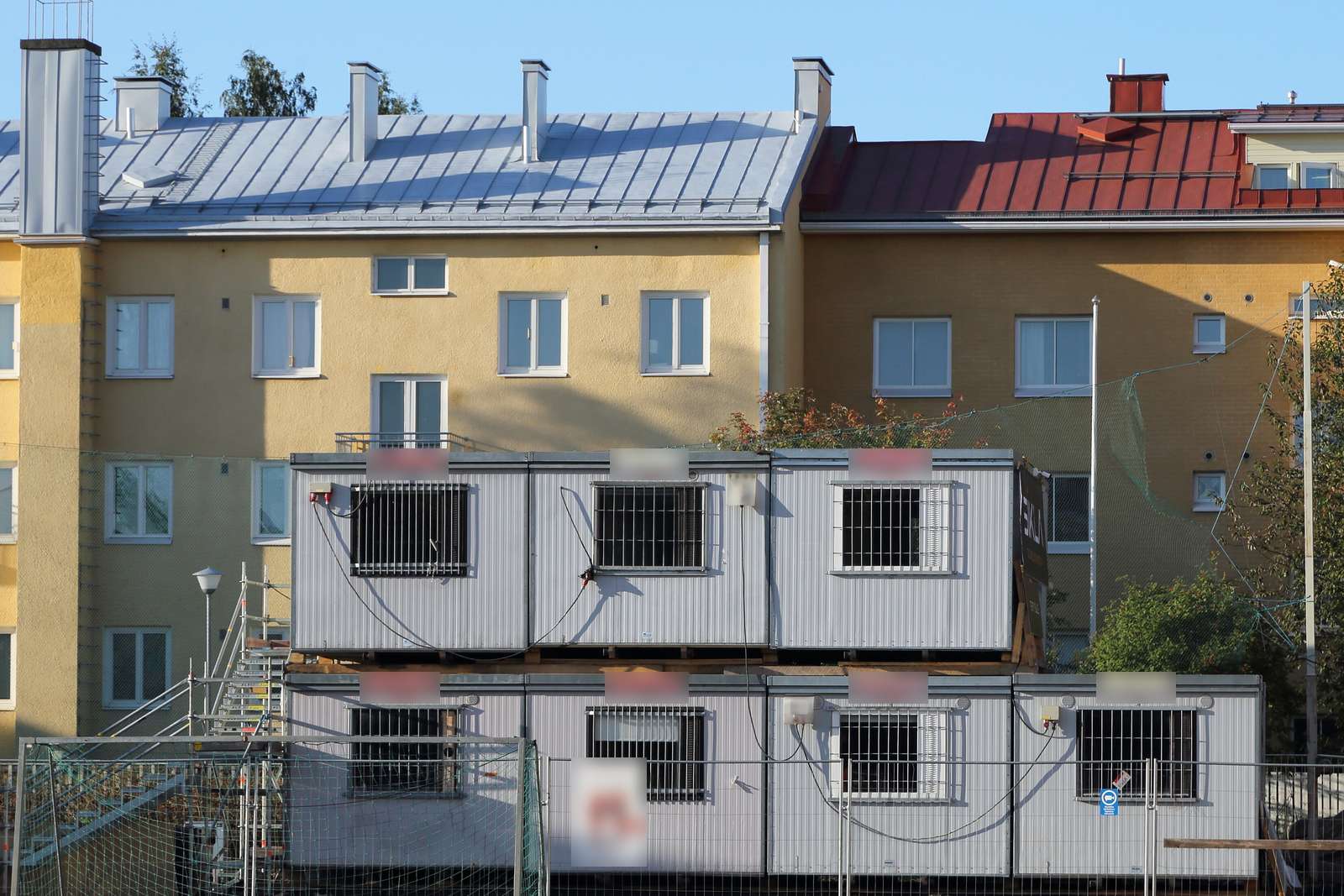 Site office containers stacked in two levels, grey with barred windows, behind protective fencing, in front of yellow apartment buildings with metal roofs