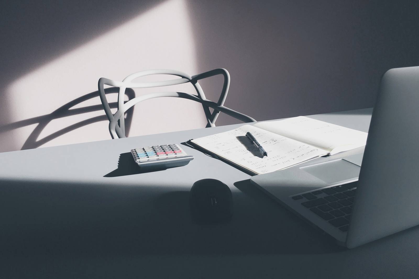 Mesa de escritório com calculadora, caderno aberto com anotações, caneta e notebook, sob luz lateral