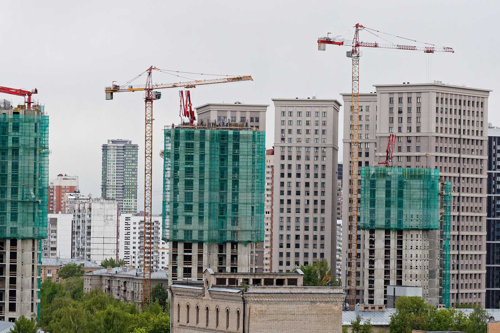 Torres residenciais em construção, duas com estrutura de concreto aparente e tela verde de proteção nos andares de cima, entre guindastes de torre amarelos e vermelhos, com prédios prontos ao fundo