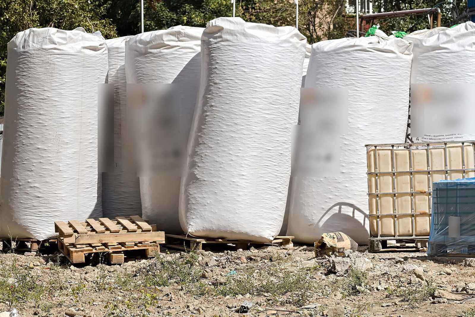 Grandes sacos de árido de pie sobre palés de madera en un terreno de obra, junto a un tanque plástico en jaula metálica y palés envueltos en film plástico